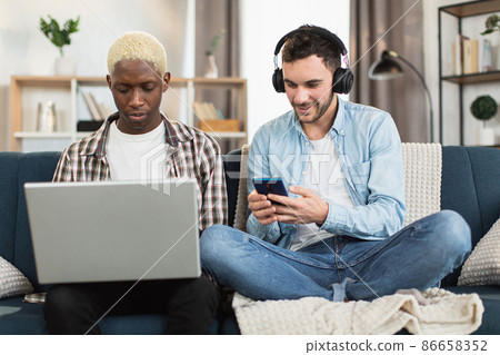 Multiracial same sex couple resting on couch with gadgets Multiracial same sex couple resting on couch with gadgets 86658352