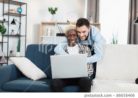 Happy same sex couple sitting on couch and using laptop Happy same sex couple sitting on couch and using laptop 86658406