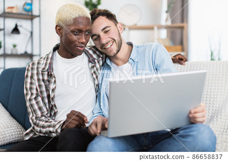 Happy couple of multiracial gays using laptop on couch 86658837