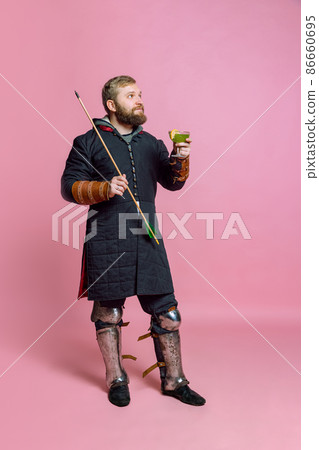 Portrait of young bearded man, medieval warrior or archer in protective cloth with arrow rising cocktail isolated over pink studio background 86660695