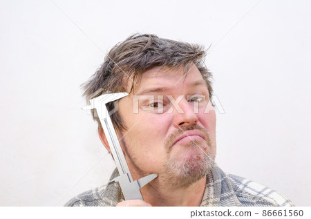 The male grimaces, measuring his face with a caliper. The male grimaces, measuring his face with a caliper. 86661560