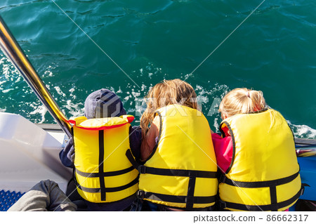 Back view three cute little friend happy excited caucasian children wear lifevest enjoy sailing on edge board motor boat sea against blue water bright sun. Summer travel vacation recreation 86662317