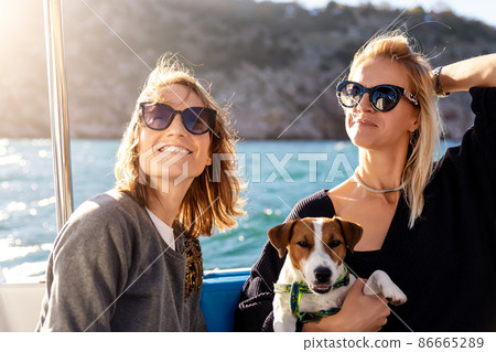 Portrait two young adult beautiful pretty caucasian woman enjoy sailing family cute adorable little jack russel dog on yacht boat against blue water bright summer day. Travel sea tourism with pets Portrait two young adult beautiful pretty caucasian woman enjoy sailing family cute adorable little jack russel dog on yacht boat against blue water bright summer day. Travel sea tourism with pets 86665289
