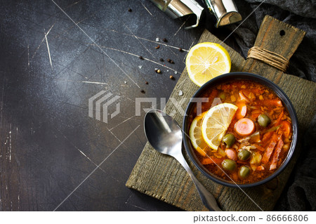 Traditional Russian soup Solyanka with meat, sausages, vegetables, capers, pickles and olives with lemon on black background. Rustic style. Top view flat lay background. Free space for your text. Traditional Russian soup Solyanka with meat, sausages, vegetables, capers, pickles and olives with lemon on black background. Rustic style. Top view flat lay background. Free space for your text. 86666806