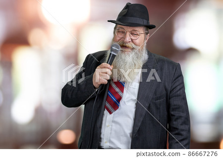Cheerful senior showman with microphone on abstract bokeh background. 86667276