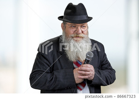 Confident senior man with beard holding microphone on blurred background. 86667323
