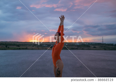 Beautiful fit caucasian girl with pink hair in red body suit posing in desert with lake on the sunset. Hands raised up. Blue sky with pink clouds. 86668519