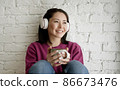 Mixed race woman holding cup of tea or coffee drinking warm beverage at home. Young asian girl in headphones listening chill music with morning drink looking through window enjoying and dreaming 86673476