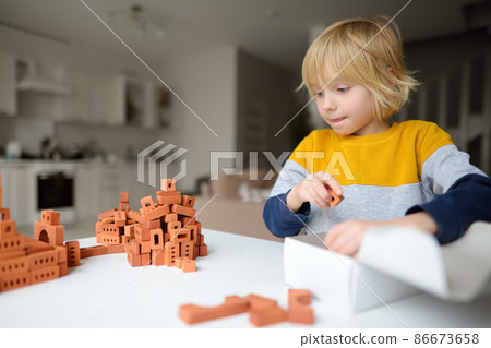 Preschooler boy is playing with real small clay bricks at the table at home. Child having fun and building smart constructions. Educational toys for kids. 86673658