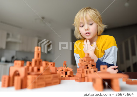Preschooler boy is playing with real small clay bricks at the table at home. Child having fun and building smart constructions. Educational toys for kids. 86673664