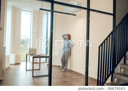 Tired woman standing leaning against wall in office Tired woman standing leaning against wall in office 86679163