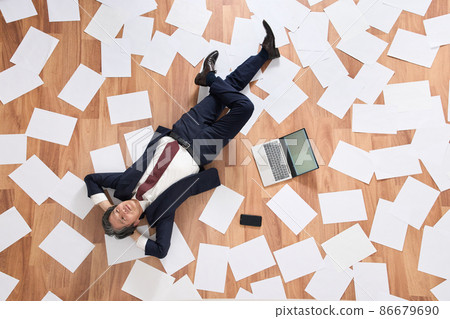 A business person in a suit lying down in a room with scattered documents A business person in a suit lying down in a room with scattered documents 86679690