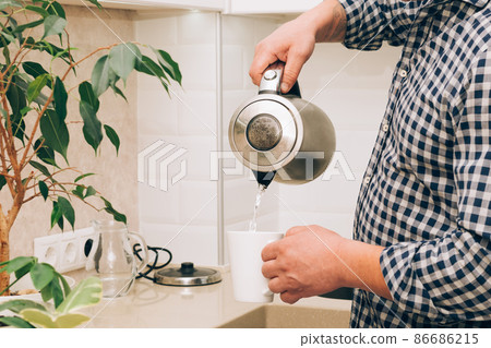A man pours hot water from a kettle into a white mug to make tea or coffee. Positive lifestyle A man pours hot water from a kettle into a white mug to make tea or coffee. Positive lifestyle 86686215