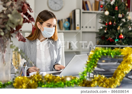 Businesswoman in mask doing her daily work in office during Christmastime Businesswoman in mask doing her daily work in office during Christmastime 86688654