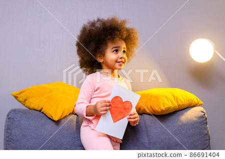 multicultural cute little curly girl holding post card with drawn heart in cozy living room at evening multicultural cute little curly girl holding post card with drawn heart in cozy living room at evening 86691404