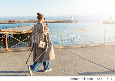 Young adult trendy stylish beautiful caucasian happy smiling woman enjoy walking by Yalta sea embankment on warm sunny day. Female person portrait wear jeans biege trench coat on urban city street Young adult trendy stylish beautiful caucasian happy smiling woman enjoy walking by Yalta sea embankment on warm sunny day. Female person portrait wear jeans biege trench coat on urban city street 86699305