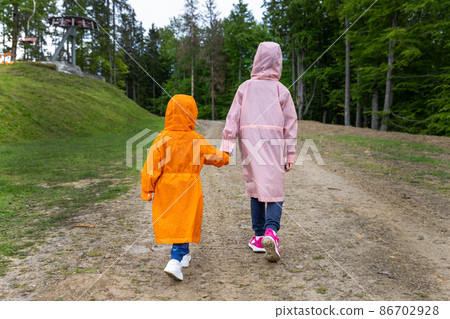 Pair of cute little caucasian sibling kids in bright waterproof raincoats walking under rain in scenic moody dirt forest road outside. Autumn weather outdoor hiking. Children friendship and support 86702928