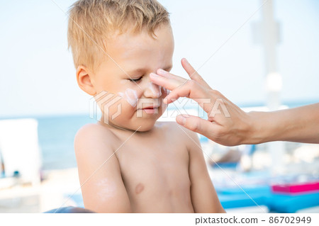 Mother applying sunscreen protection creme on cute little baby boy kid face. Mum using sunblocking lotion to protect son from sun during summer sea vacation. Child healthcare travel vacation time Mother applying sunscreen protection creme on cute little baby boy kid face. Mum using sunblocking lotion to protect son from sun during summer sea vacation. Child healthcare travel vacation time 86702949