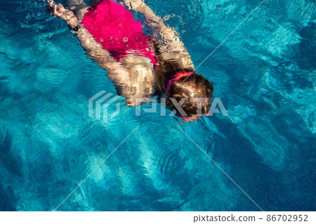 Above top view of little cute kid girl in sport pink swimsuit and goggles diving underwater in clear blue water of indoor swimming pool at dark evening time. Children watersport healthy activities 86702952