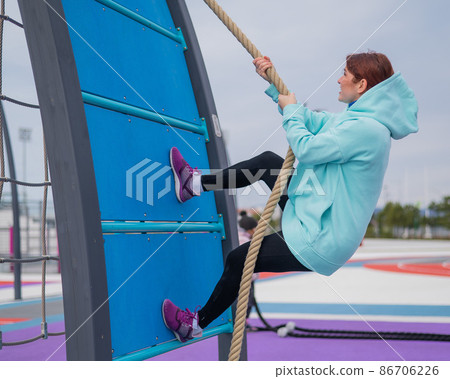 Caucasian woman in a mint sweatshirt climbing a tightrope at an outdoor sports ground.  86706226