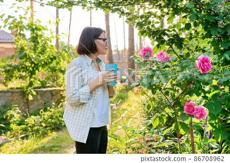 Middle-aged happy woman relaxing in garden with a cup in her hands, enjoying blooming rose Middle-aged happy woman relaxing in garden with a cup in her hands, enjoying blooming rose 86708962