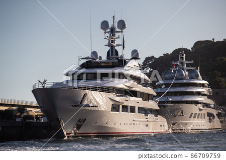 Two huge yachts are in port of Monaco at sunset, sun reflection are on glossy boards of motor boats, the chrome plated handrail, megayacht is moored in marina, mountain is on background Two huge yachts are in port of Monaco at sunset, sun reflection are on glossy boards of motor boats, the chrome plated handrail, megayacht is moored in marina, mountain is on background 86709759