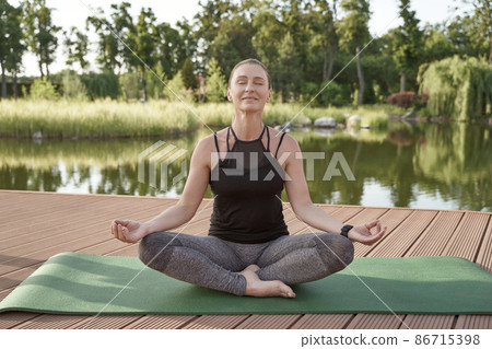 Meditation in nature. Beautiful happy woman in sportswear meditating in the morning, sitting in lotus pose on a mat with lake and green trees on background 86715398