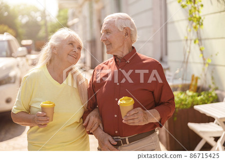 Positive senior family couple with cups walks together along street Positive senior family couple with cups walks together along street 86715425