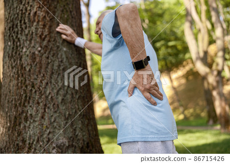 Senior man suffers from pain in lower back leaning onto tree at training in park Senior man suffers from pain in lower back leaning onto tree at training in park 86715426