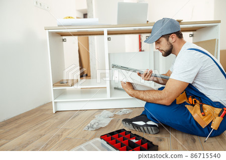 Busy handyman in overalls sitting on the floor and reading manual while assembling kitchen cabinet in apartment Busy handyman in overalls sitting on the floor and reading manual while assembling kitchen cabinet in apartment 86715540