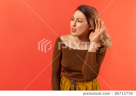 I can't hear. Portrait of pretty woman in brown blouse holding hand near ear, listening attentively with interest private conversation, confidential talk. Indoor studio shot isolated on red background I can't hear. Portrait of pretty woman in brown blouse holding hand near ear, listening attentively with interest private conversation, confidential talk. Indoor studio shot isolated on red background 86715750