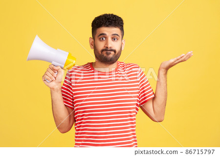 Puzzled confused man with beard in striped t-shirt shrugging shoulders holding in hands megaphone, cant find common language, communication problems. Indoor studio shot isolated on yellow background Puzzled confused man with beard in striped t-shirt shrugging shoulders holding in hands megaphone, cant find common language, communication problems. Indoor studio shot isolated on yellow background 86715797