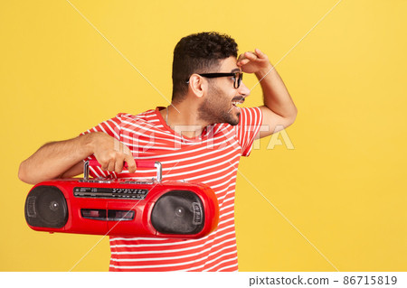 Happy bearded man in eyeglasses and striped t-shirt looking far away holding hand above forehead and holding retro tape recorder, waiting for friends. Indoor studio shot isolated on yellow background 86715819