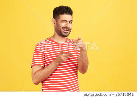 Excited mischievous man with beard in striped t-shirt winking looking and pointing fingers at camera, coquetting and flirting. Indoor studio shot isolated on yellow background Excited mischievous man with beard in striped t-shirt winking looking and pointing fingers at camera, coquetting and flirting. Indoor studio shot isolated on yellow background 86715841