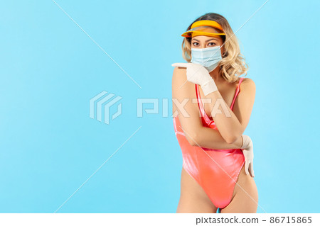 Look info about covid-19. Woman in swimsuit, wearing hygienic face mask and surgical gloves to prevent coronavirus on resort, showing blank advertising area, summer vacation with covid-19. studio shot 86715865