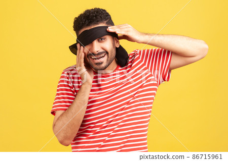 Happy bearded man in striped red t-shirt taking off blindfold from eyes to look at his surprise, looking at camera with toothy smile. Indoor studio shot isolated on yellow background 86715961