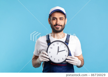Courier delivery on time. Portrait of positive bearded handyman in overalls and gloves holding clock, smiling to camera. Professional and punctual service of house repair, maintenance. studio shot 86716029