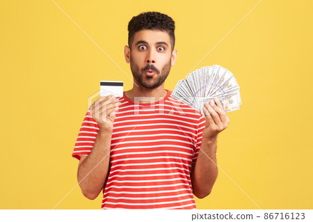 Shocked bearded man in striped t-shirt holding and showing dollar cash and credit card, surprised with cashback, currency exchange. Indoor studio shot isolated on yellow background 86716123