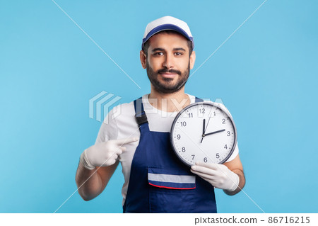 Courier delivery on time. Optimistic positive bearded handyman in overalls and gloves pointing clock, smiling to camera. Professional and punctual service of house repair, maintenance. studio shot 86716215