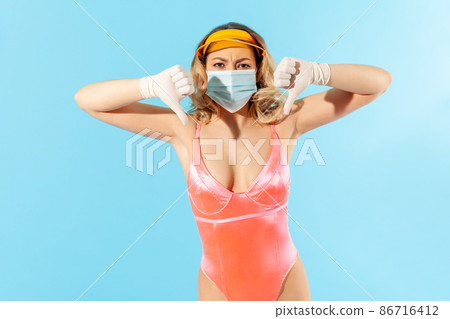 Displeased woman in swimsuit, wearing hygienic face mask and surgical gloves to prevent contagious coronavirus on resort, showing thumbs down, dislike, screw worst summer with covid-19. studio shot 86716412