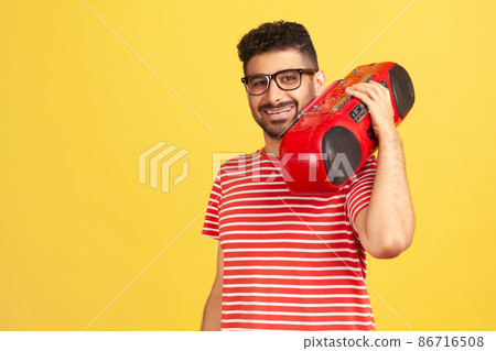 Positive bearded man in eyeglasses and striped t-shirt listening to music holding red tape recorder on shoulder, looking at camera with toothy smile. Indoor studio shot isolated on yellow background 86716508