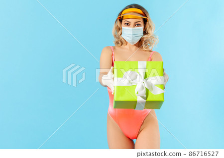 Take this gift. Woman in swimsuit offering wrapped box to camera, wearing hygienic face mask and gloves to give present safely, prevent coronavirus covid-19 infection. studio shot, isolated advertise Take this gift. Woman in swimsuit offering wrapped box to camera, wearing hygienic face mask and gloves to give present safely, prevent coronavirus covid-19 infection. studio shot, isolated advertise 86716527