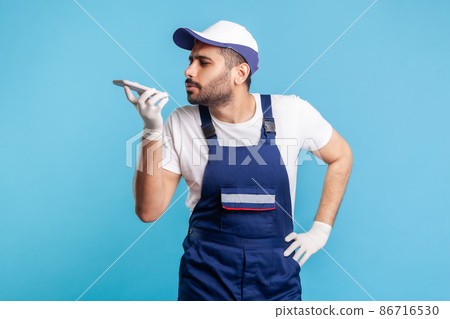 Virtual assistant. Handyman in overalls and gloves talking to phone recording message, using digital voice app to accept online order as delivery, repair and maintenance services. studio shot isolated 86716530