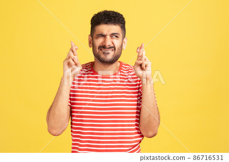 Funny hopeful man with beard in striped t-shirt crossing fingers for luck, making a wish, dreaming of innermost, ritual. Indoor studio shot isolated on yellow background Funny hopeful man with beard in striped t-shirt crossing fingers for luck, making a wish, dreaming of innermost, ritual. Indoor studio shot isolated on yellow background 86716531
