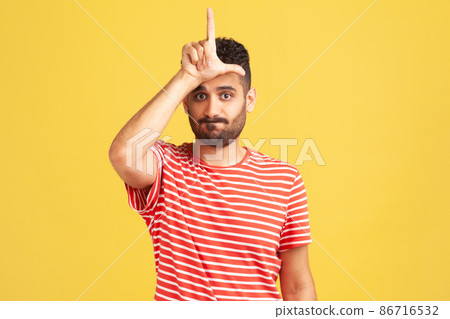 Gloomy depressed man with beard in striped t-shirt showing looser gesture holding fingers near forehead, sad because of silly mistake. Indoor studio shot isolated on yellow background 86716532