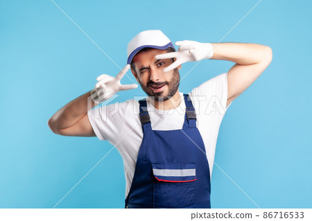 Positive handyman in overalls and hygiene gloves showing victory sign and winking playfully to camera, profession of service industry, courier delivery, housekeeping maintenance. studio shot isolated Positive handyman in overalls and hygiene gloves showing victory sign and winking playfully to camera, profession of service industry, courier delivery, housekeeping maintenance. studio shot isolated 86716533