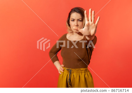 Stop, there is no way. Portrait of serious young woman with hand on the waist in casual clothes demonstrate stop gesture by her hand. Forbidden and decline concept. Indoor isolated on red background 86716714