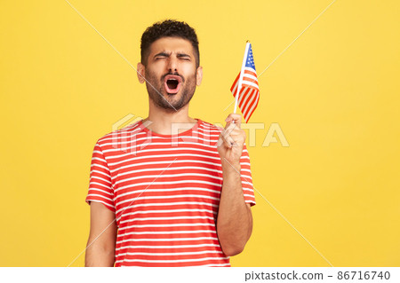 Devoted patriotic man in striped t-shirt holding in hand flag of united states of america, singing hymn celebrating independence day. Indoor studio shot isolated on yellow background 86716740