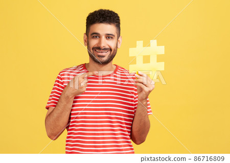 Positive bearded man blogger in striped t-shirt pointing at yellow hashtag symbol in his hand, recommending posts, looking at camera with toothy smile. Indoor studio shot isolated on yellow background 86716809