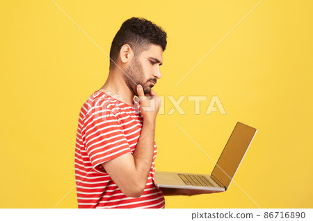 Profile portrait thoughtful pensive man with beard in striped t-shirt holding laptop and rubbing chin looking at display, thinking over new business project. Indoor isolated on yellow background 86716890